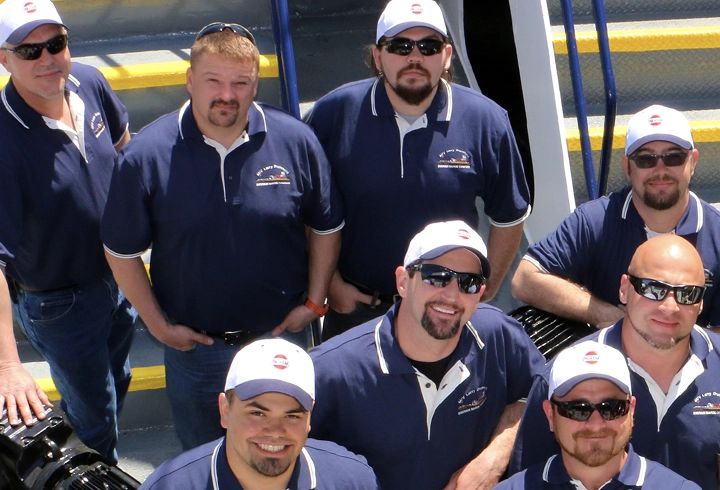 Eight men wearing matching blue polos, white caps, and sunglasses pose together near a set of stairs outdoors.