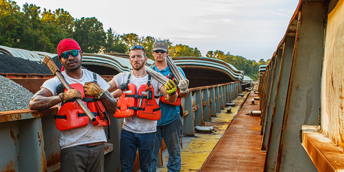 Three workers wearing life vests and holding tools stand on a barge with piles of gravel and trees in the background.