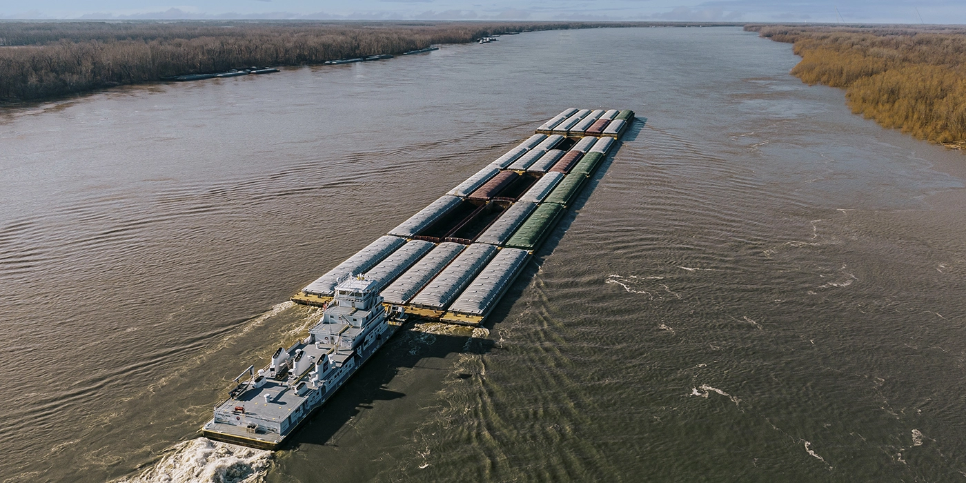 A towboat pushing a long row of barges up a wide river, with trees lining both riverbanks.