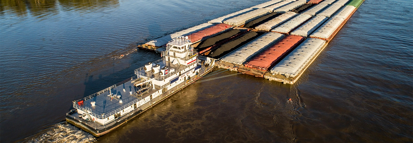 A towboat pushes multiple covered barges along a wide river, viewed from above.
