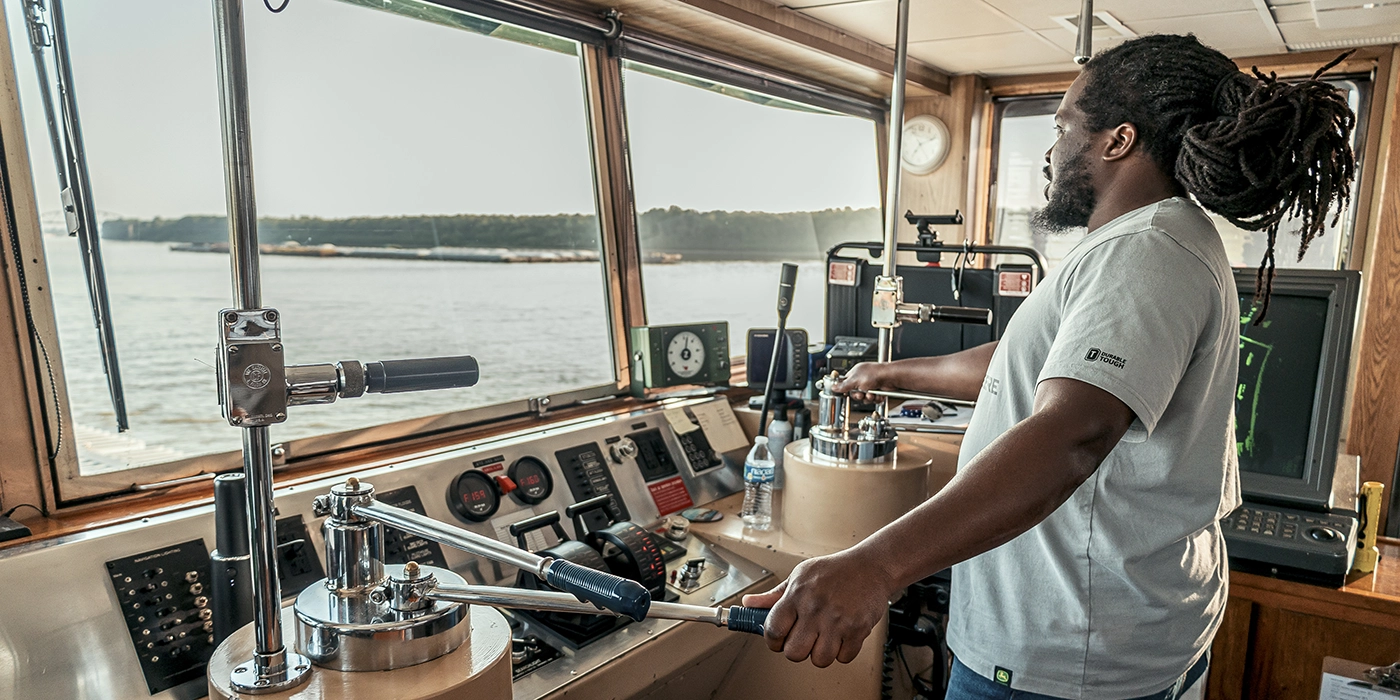 Person operating the controls inside a boat or ships wheelhouse, looking out toward the water through large windows.