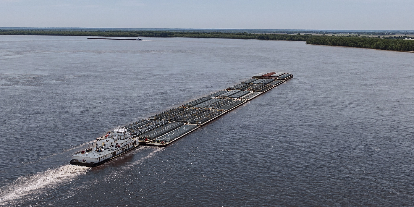 A barge pushes multiple cargo containers down a wide river with trees lining the distant shoreline.
