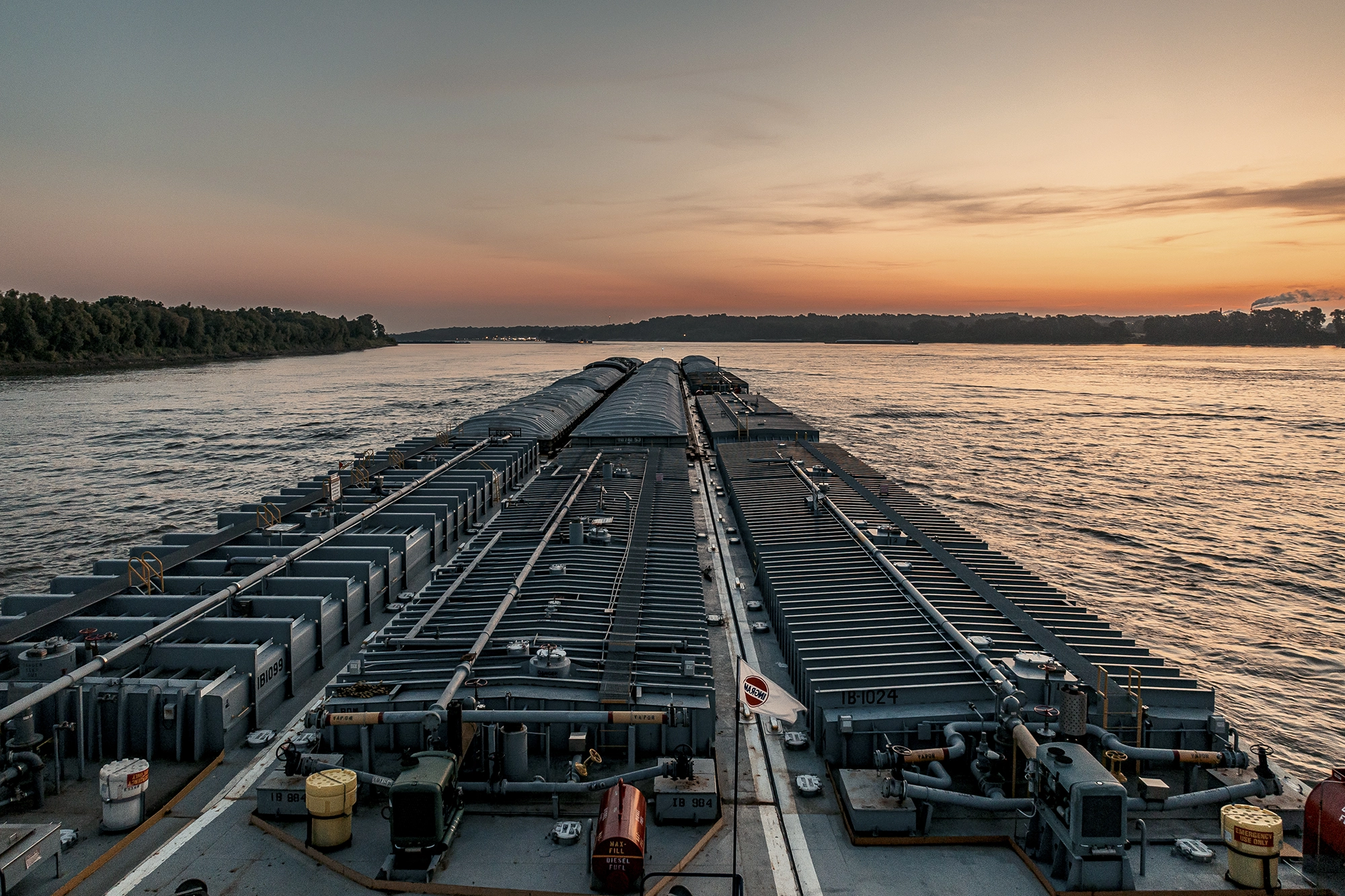 A large cargo barge travels down a wide river at sunset with trees lining the distant shore.