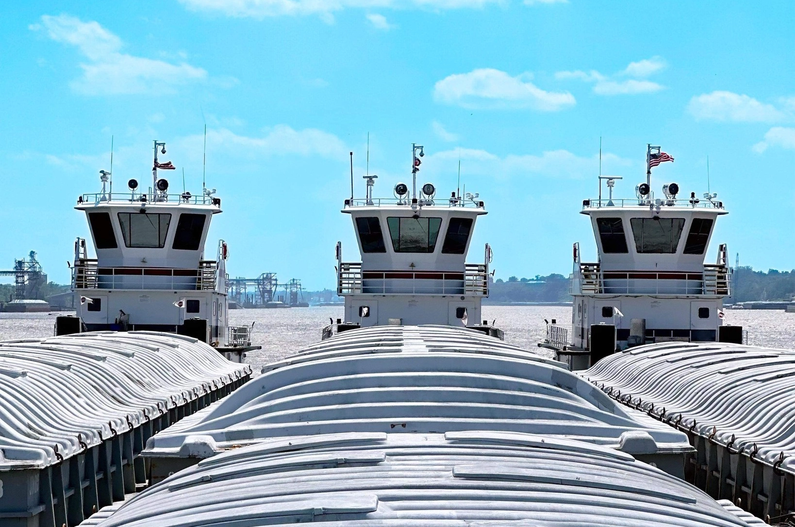 Three towboats are lined up on a river, with barges in the foreground and an industrial shoreline in the background.