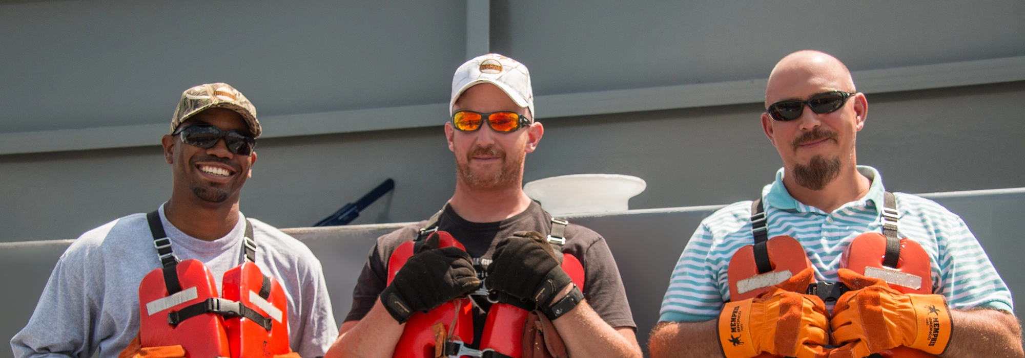 Three men wearing life vests, gloves, and sunglasses stand side by side on a boat.