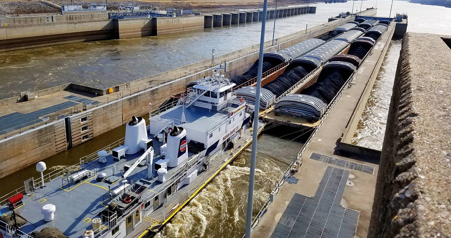 A towboat pushes barges carrying bulk cargo through a lock on a river next to a dam structure.